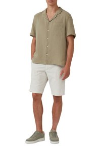 Kurzärmliges, olivgrünes Hemd mit strukturierter Oberfläche, Knopfleiste und Brusttasche, kombiniert mit weißen Shorts und hellgrünen Sneakern.