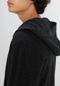 Dunkelgrauer Hoodie mit weichem Material, lockerer Passform und großer Kapuze mit doppellagigem Rand. Sichtbare Nähte verleihen Details.