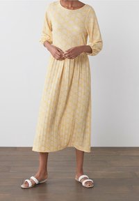 Vestido midi amarillo con escote redondo y mangas tres cuartos, que presenta un patrón texturizado de círculos blancos. Combinado con sandalias trenzadas blancas.