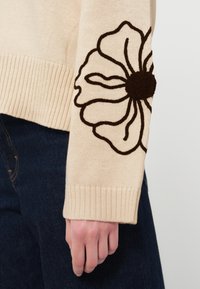 Béžový svetr s žebrovanými manžetami, na levém rukávu velký tmavě hnědý vyšívaný květinový motiv. Měkká pletená textura.