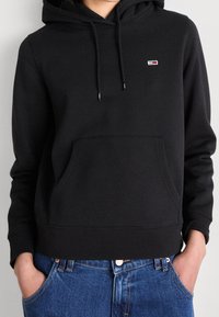 Sudadera negra de tela suave, con un bolsillo delantero, capucha con cordón y un pequeño logo bordado en el pecho. Llevada con jeans azules.