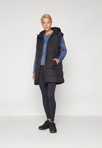 Juodas pūkinis liemenė su gaubtu, vertikaliomis siūlėmis, dviem šoninėmis kišenėmis ir užtrauktuku priekyje. Derinamas su mėlynu ilgomis rankovėmis viršutiniu drabužiu ir juodomis tamprėmis.