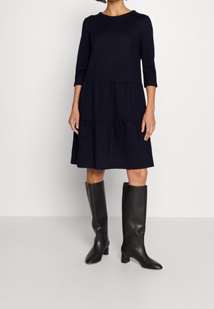 Kobieta w granatowej, warstwowej sukience midi z rękawami do łokcia oraz czarnych, wysokich botkach na obcasie, stojąca na tle jednolitego tła.
