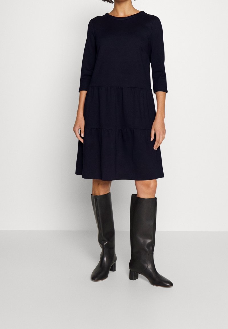 Femme portant une robe bleu marine à volants jusqu'aux genoux avec des manches trois-quarts et des bottes à talons noires jusqu'aux genoux, debout devant un fond uni.