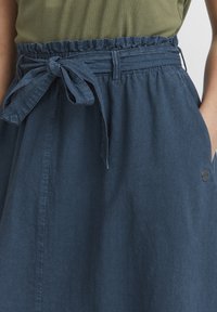 Jupe bleu foncé avec une finition texturée, taille élastique et un détail de nœud à l'avant. Comprend des poches latérales et un petit accent en métal.