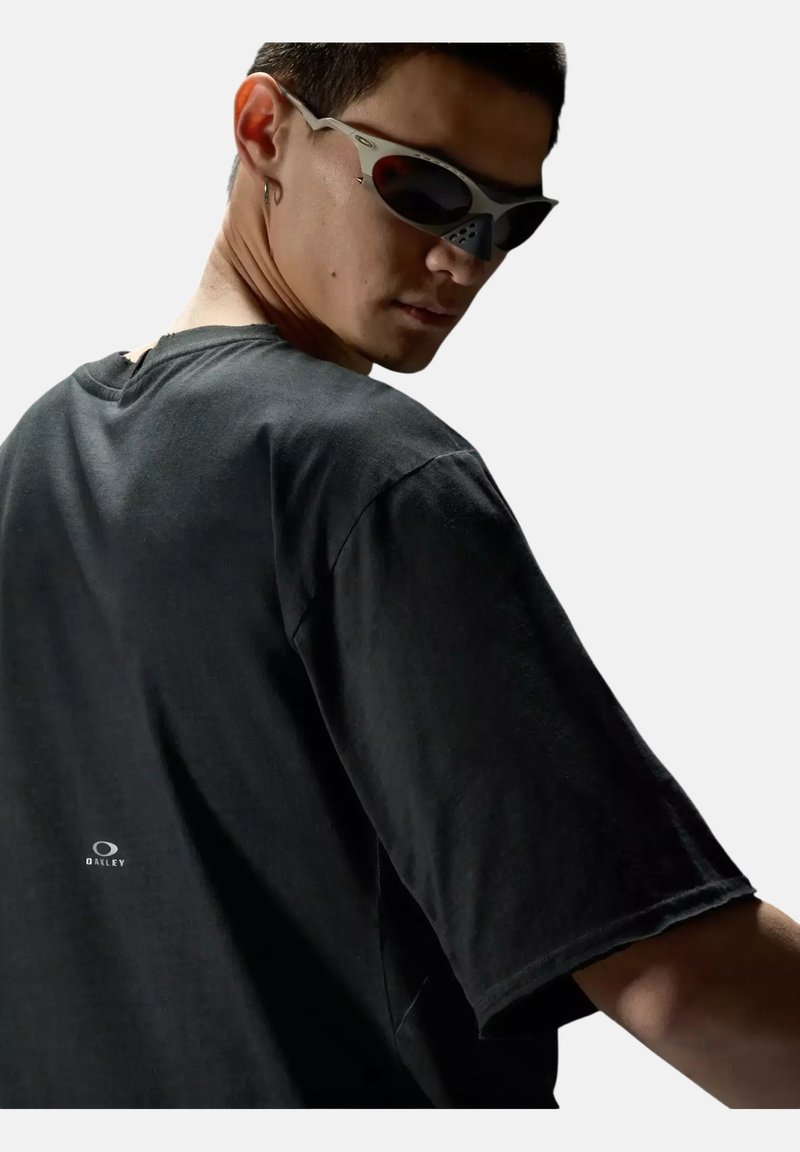 Jeune homme portant un t-shirt noir Oakley et des lunettes de soleil futuristes grises, regardant par-dessus son épaule sur fond blanc.