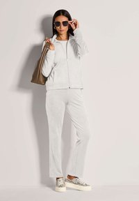 Vrouw in een lichtgrijze gewatteerde jas en broek, met een bruine leren handtas, die grote donkere zonnebril aanpast, staande tegen een effen muur.