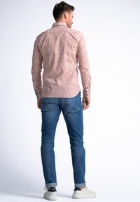 Camisa de manga larga en rosa claro con patrón floral blanco. Presenta un cuello tipo polo y un dobladillo redondeado. Combinada con jeans azules y zapatos blancos.