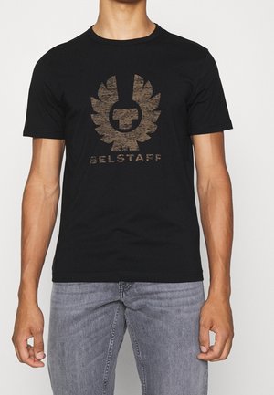 Mann trägt ein schwarzes Kurzarm-T-Shirt mit einem großen braunen Belstaff-Logo und Text auf der Vorderseite, kombiniert mit hellgrauen Jeans.