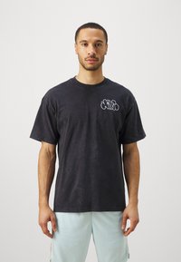 Hombre de pie contra un fondo blanco, vistiendo una camiseta negra de gran tamaño con un logo blanco en el pecho y pantalones cortos azul claro.