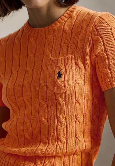 Άτομο που φοράει κοντομάνικη, πορτοκαλί πουλόβερ με πλέξη καλώδιο και μια μικρή τσέπη με σκούρο κεντητό λογότυπο στο στήθος.