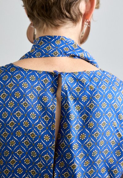 Blaue gemusterte Stoff mit floralen Designs, ausgestattet mit einem offenen Rücken und einer Bindeschleife am Hals. Glatte Textur und leichtes Material.