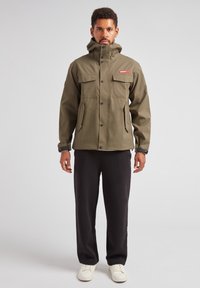 Giacca impermeabile verde oliva con colletto alto, bottoni a pressione, due tasche sul petto e polsini regolabili. Indossata con pantaloni scuri e scarpe bianche.