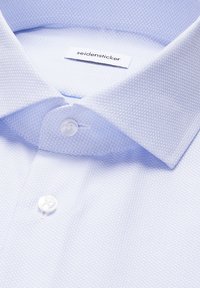 Gros plan sur le col d'une chemise habillée texturée bleu clair avec des boutons blancs et une étiquette de marque "seidensticker" visible à l'intérieur du col.