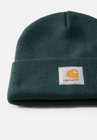 Mørkegrønn ribbestrikket lue med brettet kant og hvit firkantet Carhartt-logo-kommer med oransje emblem og rødt tekst.