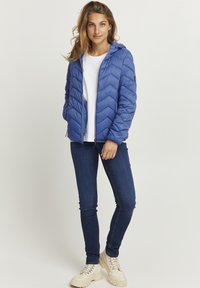 Doudoune bleue avec des coutures en chevron, poches zippées et capuche, portée avec un t-shirt blanc et un jean foncé, complétée par des chaussures beiges.