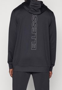 Personne portant un sweat à capuche noir avec le logo vertical blanc "ELLESSE" dans le dos, associé à un pantalon noir assorti.
