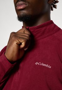Bordeaux fleece genser med glidelås-krage og brodert "Columbia" logo. Har myk tekstur og tettsittende mansjetter.