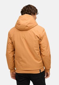 Uomo con giacca con cappuccio color sabbia e pantaloni neri, visto di spalle su uno sfondo chiaro e uniforme.