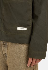 Olivgrüne Jacke aus strukturiertem Material mit einer Brusttasche. Verfügt über ein weißes Etikett mit der Aufschrift "ELEMENT CO. SINCE 1992" am unteren Rand.