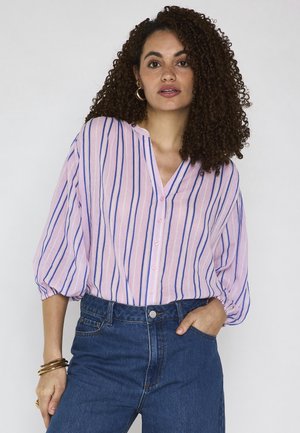 Donna con capelli ricci che indossa una camicetta a righe rosa e blu e jeans blu, posando con una mano in tasca e un'espressione neutra.