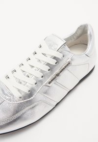 Sneaker argenté avec une finition en cuir métallique, lacets blancs et un design épuré. Présente un logo sur le côté et une semelle en caoutchouc noir.