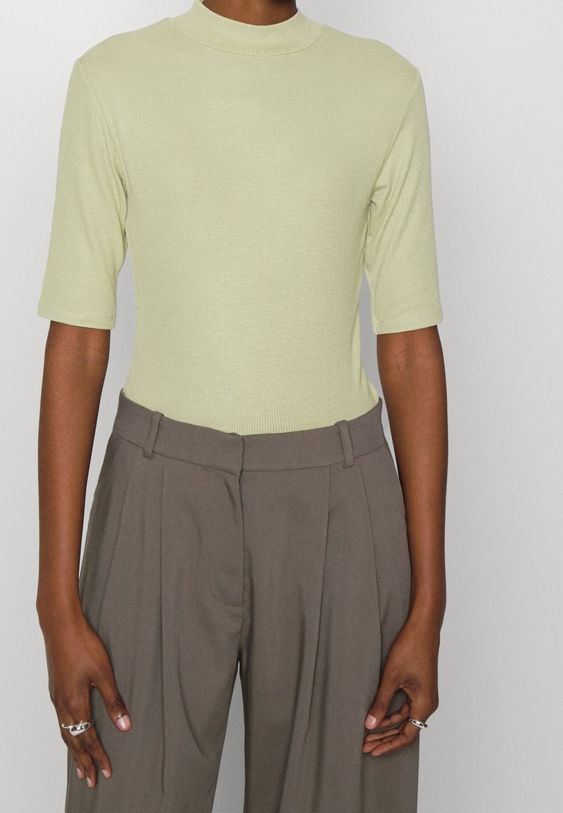 Personne portant un haut côtelé vert clair à manches courtes rentré dans un pantalon marron ample taille haute avec passants de ceinture.