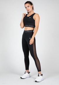 Black sports bra with logo band, paired with high-waisted leggings featuring sheer side panels. White athletic shoes complete the look.
