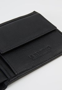 Portofel din piele neagră cu un finisaj texturat. Prezintă un logo embosat și un sistem de închidere cu clapetă. Design compact cu detalii de cusătură vizibile.