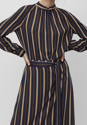 Kobieta w granatowej sukience z pionowymi białymi i musztardowymi paskami, długimi rękawami, wysokim kołnierzem i związanym paskiem w talii.