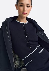 Soe tumesinistest niitidest kampsun valge lainelise kaeluse ja nuppudega, mis on kaunistatud kunsthelmestega; kihiliselt kantud tumeda jaki all, millel on ruuduline vooder.