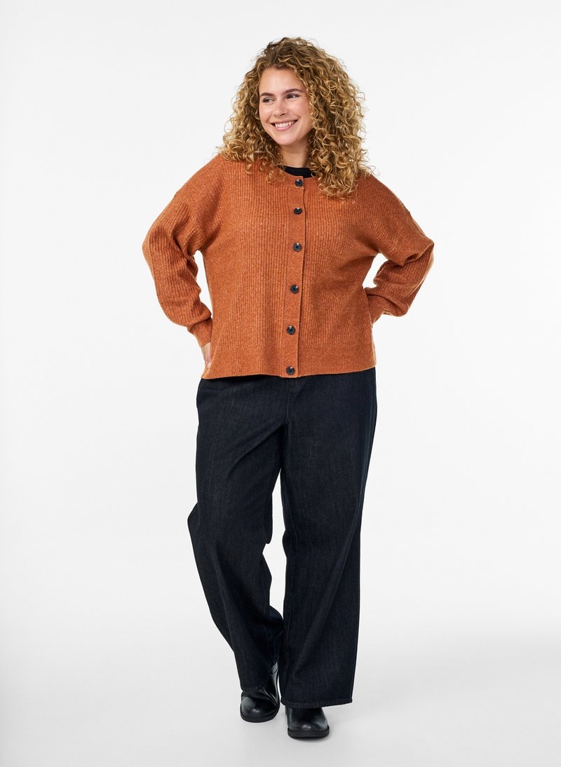 Mujer de cabello rizado sonriendo, vistiendo un cárdigan abotonado color óxido, pantalones negros de pierna ancha y zapatos negros, de pie con las manos en las caderas.