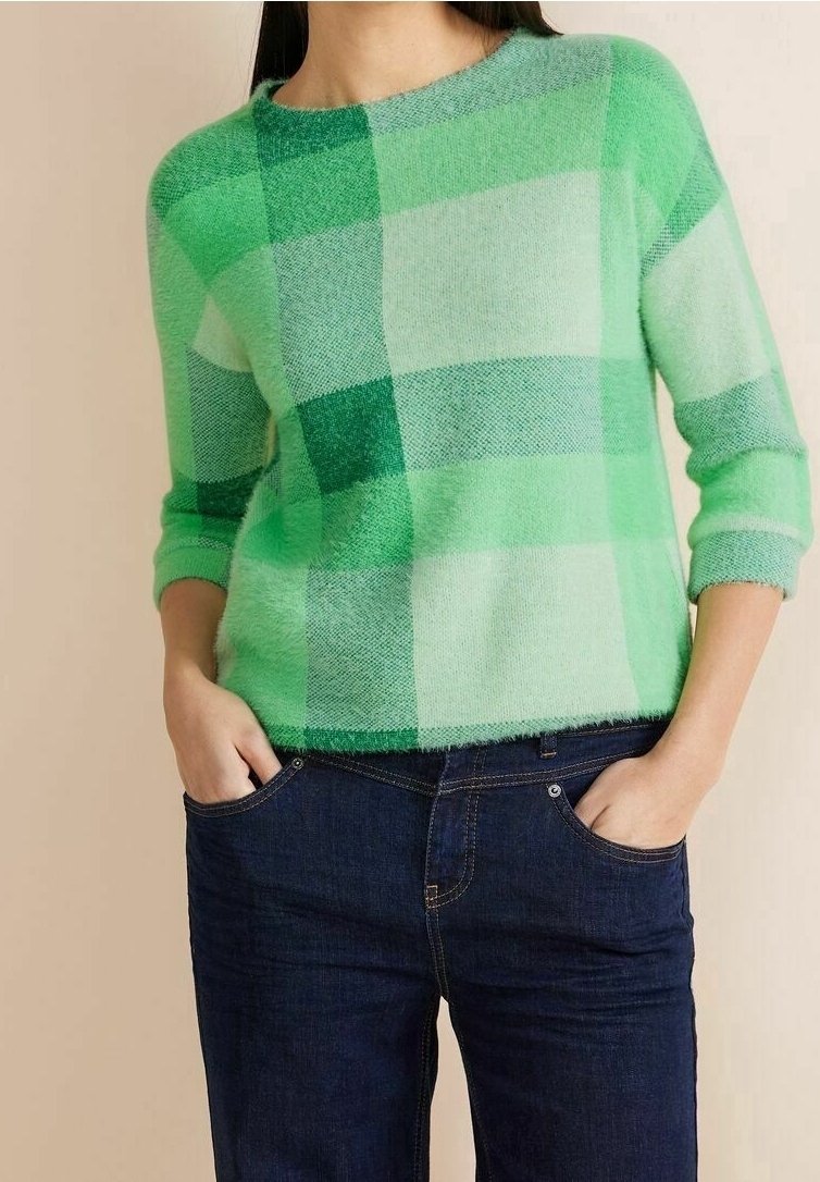 Femme portant un pull à carreaux vert avec les manches retroussées et un jean bleu foncé, mains dans les poches, debout devant un fond neutre.