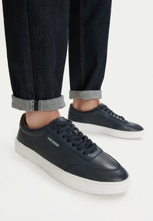 Czarne skórzane sneakersy z białą podeszwą noszone z podwiniętymi ciemnymi jeansami.