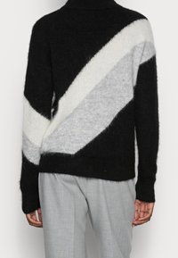 Pull en maille noire avec des rayures diagonales crème et gris clair. Il comporte un col montant et un ourlet côtelé, avec une texture douce et duveteuse.