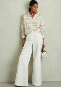 Lys beige stribet sweater med krave, parret med højtaljede hvide vidde bukser. Modellen holder en lille brun og cremefarvet taske.
