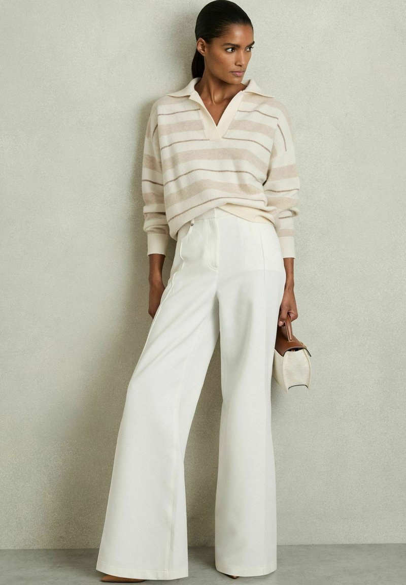 Lys beige stribet sweater med krave, parret med højtaljede hvide vidde bukser. Modellen holder en lille brun og cremefarvet taske.