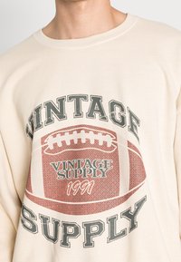 Béžová mikina s velkým grafickým potiskem ve vintage stylu s motivem fotbalu a textem "VINTAGE SUPPLY 1991" vytištěným na přední straně.