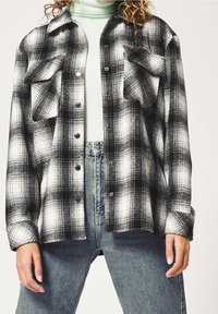 Svart och vit rutigt flanellskjorta med knappstängning fram, två bröstfickor och avslappnad passform. Bärs över en ljus mintgrön polotröja.