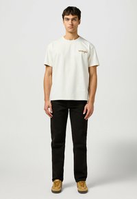 Camiseta de manga corta color beige claro con el logotipo "Wrangler" en dorado, combinada con jeans negros y zapatillas color crema, creando un look casual.