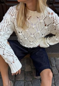 Kreemi värvi kudum, millel on pitsimuster ja soonilised varrukad, koos tumedate siniste teksashortsidega. Nähtavad on tekstuurilised ja avatud silmuste detailid.