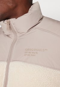 Beige zip-up-jacka med en texturerad ljuscremefleecepanel. Har broderad text, hög krage och slät yttermaterial.