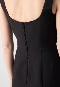 Black dress featuring a scoop back, structured fit, and a row of textured buttons along the back seam. Smooth fabric with no visible patterns.