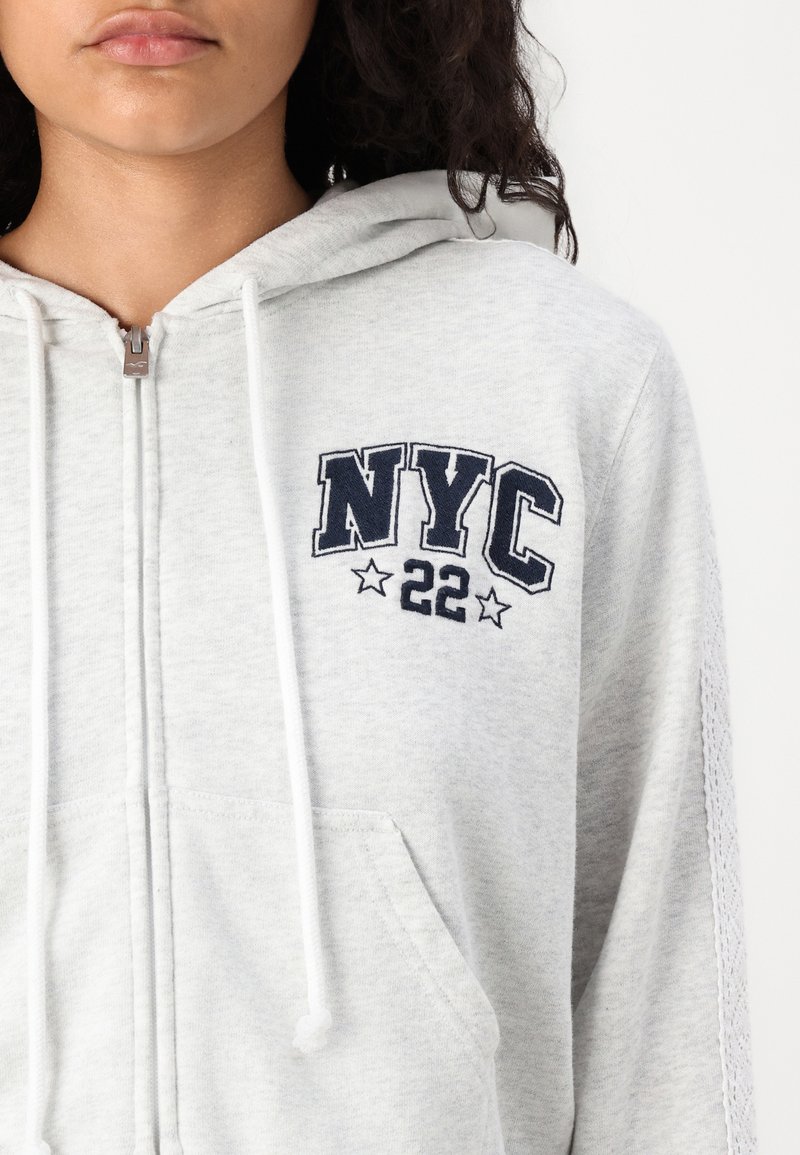 Felpa grigia con zip e drawstring, con la scritta "NYC 22" ricamata in blu marino, tasche frontali e dettagli laterali testurizzati.