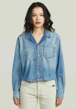 Frau mit welligem dunklem Haar trägt eine hellblaue, kurz geschnittene Jeansjacke mit Knöpfen und Tasche vorne, kombiniert mit cremeweißen Hosen.