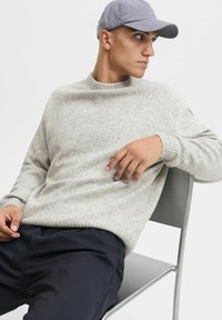 Jasnoszara, teksturowana sweter z okrągłym dekoltem, noszony na ciemnych spodniach, zestawiony z szarą czapką, siedzący na prostym metalowym krześle.