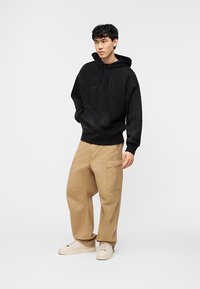 Νεαρός άνδρας που στέκεται και φοράει μαύρο φούτερ GAP, μπεζ παντελόνι cargo και εκρού αθλητικά, κοιτάζοντας προς το πλάι με τα χέρια στις τσέπες.