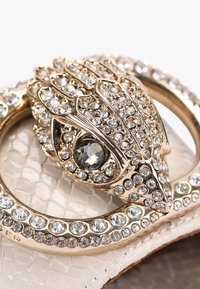 Spilla color oro con un design decorativo che ricorda una creatura stilizzata, abbellita con strass trasparenti, su uno sfondo leggero e texturizzato.