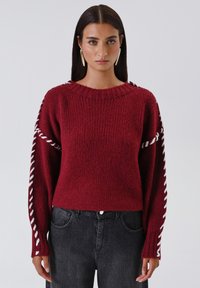 Una donna con capelli scuri lisci indossa un maglione lavorato a maglia rosso con dettagli bianchi intrecciati sulle maniche e jeans neri a vita alta.