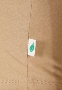 Beiger Stoff mit glattem, dehnbarem Material. Ein kleines weißes Etikett mit einem grünen Blättermuster ist an der Seitennaht angenäht.
